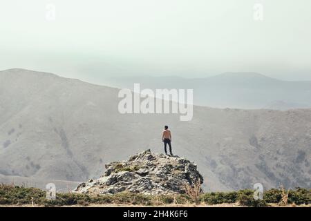 Ein junger Mann, von hinten gesehen, steht auf einem Felsen, um die Aussicht auf die Berge vor ihm zu betrachten. Stockfoto