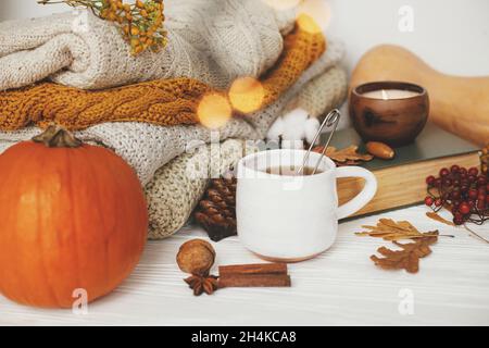 Kürbis, eine Tasse Tee, kuschelige Pullover, Herbstblätter, brennende Kerze und ein Vintage-Buch in goldenen Lichtern Bokeh auf weißem Holzhintergrund im Zimmer. Hallo au Stockfoto