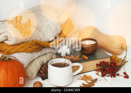Kürbis, eine Tasse Tee, kuschelige Pullover, Herbstblätter, brennende Kerze und ein Vintage-Buch in goldenen Lichtern Bokeh auf weißem Holzhintergrund im Zimmer. Hallo au Stockfoto