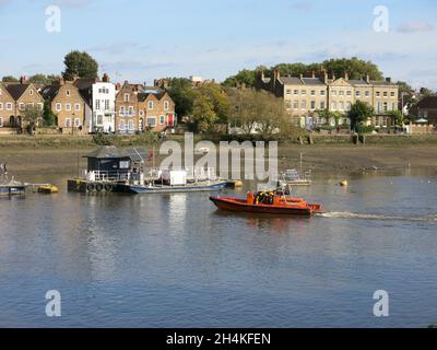 Blick vom Thames Path nach Norden über den Fluss in Richtung Gebäude entlang des Strand-on-the-Green, Chiswick, mit Booten, die an schwimmenden Liegeplätzen festgemacht sind. Stockfoto