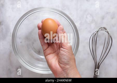 Draufsicht auf die rechte Frauenhand, die ein rohes braunes Ei über der Glasschale hält und auf der Marmoroberfläche Schneebesen Stockfoto