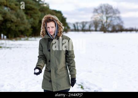 Ein entschlossen aussehender junger Mann mit einem Schneeball in der Hand, der bereit ist, ihn während einer Schneeballschlacht zu werfen. Er ist in einer schneebedeckten winterlichen Landschaft Stockfoto