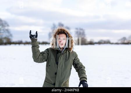 Ein entschlossen aussehender junger Mann mit einem Schneeball in der Hand, der bereit ist, ihn während einer Schneeballschlacht zu werfen. Er ist in einer schneebedeckten winterlichen Landschaft Stockfoto