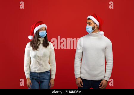 Soziale Distanzierung während der Covid-19-Sperre. Arabische Ehepartner in Santa-Hüten und Schutzmasken, die sich gegenseitig angucken, stehen auf rotem Hintergrund, leere s Stockfoto