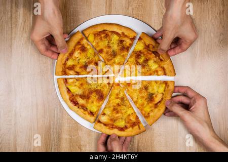 Draufsicht auf die Hände, die köstliche und knusprige hawaiianische Pizzastücke mit Mozzarella-Käse, Ananas und Tomatenquelle nehmen. Gruppe hungriger Fr. Stockfoto