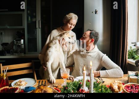 Frontansicht des glücklichen jungen verliebten Paares, das während der Weihnachtsfeier mit Akita Hund am festlichen Weihnachtstisch sitzt. Stockfoto