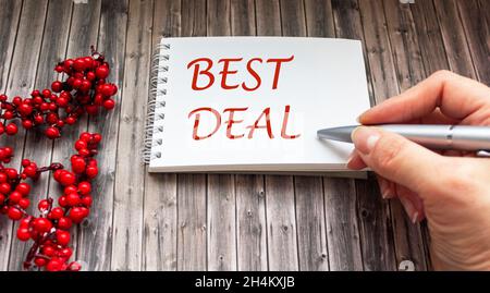Eine weibliche Hand schreibt auf ein Notizbuch den BESTEN DEAL, auf einem Holztisch liegt ein Zweig der Eberesche Stockfoto