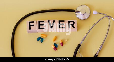 Das Wort Fever stand auf einem Holzblock. Es gibt ein Stethoskop und Pillen auf einem gelben Tisch. Stockfoto
