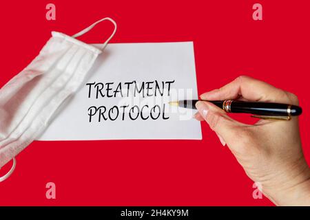 Das Wort Behandlungsprotokoll wird von einem Arzt auf einem roten Hintergrund in der Nähe einer medizinischen Schutzmaske geschrieben. Medizinisches Konzept Stockfoto