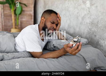 Schockierter, schwarzer Mann, der den Wecker ansah, zu Hause im Bett lag, den Kopf berührte und zu spät zur Arbeit kam. Gestresster afroamerikanischer Kerl überschlafend, Stockfoto