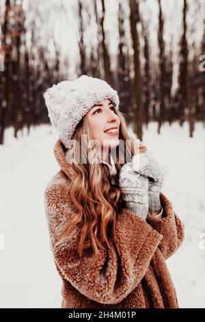 Schöne junge Frau in schneebedeckten Winterwäldern. Mädchen trägt flauschige Handschuhe, Mütze und Mantel. Weihnachtswald, Bäume auf verschwommenem Hintergrund. Gekreuzt Stockfoto