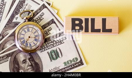 Das Wort Bill steht auf einem Holzblock, einem Stapel von 100 Dollar und einer Uhr auf gelbem Hintergrund. Amerikanische Währung. Geld- und Finanzkonzept. Stockfoto