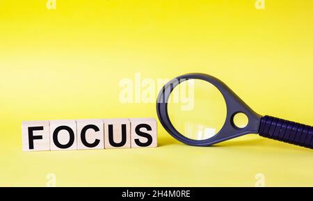 FOCUS - ein Wort aus Holzwürfeln mit schwarzen Buchstaben, auf einem gelben Hintergrund, in der Nähe einer Lupe. Stockfoto