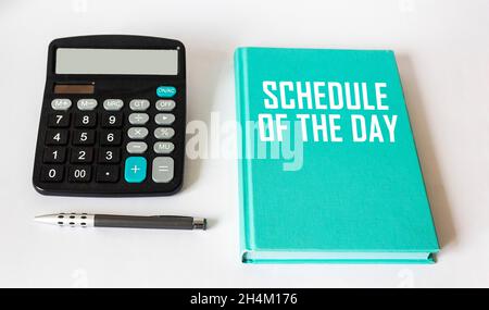 ZEITPLAN DES TAGES geschrieben auf einem grünen Notizbuch, neben einem Rechner und einem Stift auf weißem Hintergrund. Stockfoto