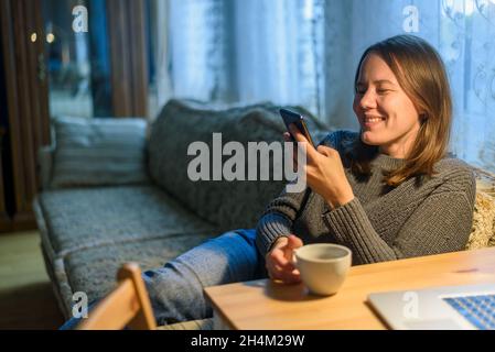 Frau im Pullover, die ihr Telefon in der Nähe des Laptops im Wohnzimmer benutzt Stockfoto
