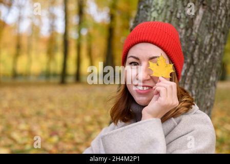 Frau in rot gestricktem Hut hält Ahornblatt in der Nähe ihres Auges im Herbstpark Stockfoto