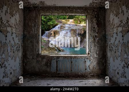 Blick auf einen tropischen Wasserfall durch ein Fensterloch in einem beschädigten verlassenen Plattenhaus. Stockfoto