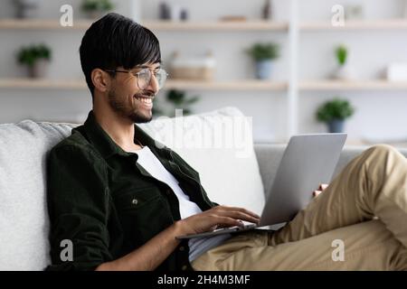 Positiver inder, der alleine zu Hause chillt, auf der Couch sitzt und einen Laptop benutzt, mit Damen auf der Dating-Website chattet, scrollt oder auf der Suche nach einem Job ist Stockfoto