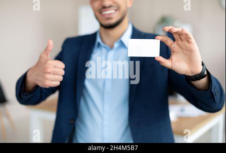 Nicht erkennbarer arabischer Unternehmer, der leere Visitenkarten hält, den Daumen im Büro hochstischt, Design-Mockup anstellt. Beschnittene Ansicht des östlichen Geschäftsmannes dem Stockfoto