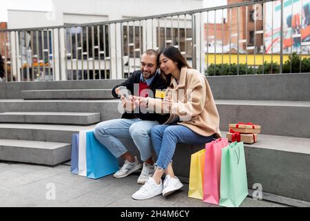 Erstellen von Online-Bestellkonzepts. Glückliches multirassisches Paar, das auf Steintreppen mit Smartphone und Kreditkarte sitzt, im Internet einkaufen, umgeben von s Stockfoto