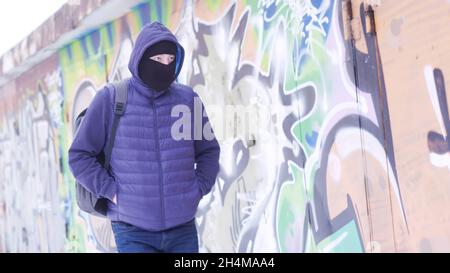 Seriöser junger Mann in lässigem Outfit und Balaclava, der entlang der Graffitti-Wand läuft. Männlich mit einem Rucksack versteckt sein Gesicht und schaut sich um, Konzept der Stockfoto