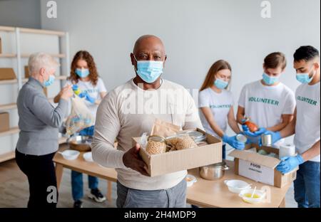 Wohltätigkeitsorganisation. afroamerikanischer Senioren hält einen Karton mit Spenden, Essen, trägt eine medizinische Maske und schaut in die Kamera. Gruppe von Freiwilligen gibt Stockfoto