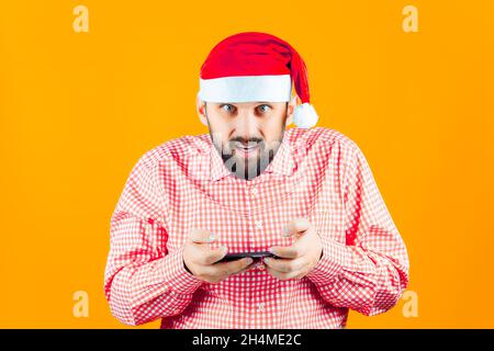 Ein Mann in einem Weihnachtsmannhut auf gelbem Hintergrund hält ein Telefon mit zwei Händen und schützt seine Schultern Stockfoto