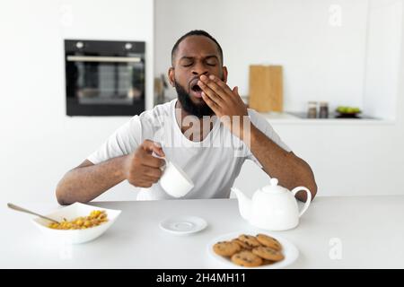 Konzept Für Fehlenden Schlaf. Porträt eines müden jungen afroamerikanischen Mannes, der am Esstisch in der Küche schläft, und eines schläfrigen Mannes, der den Coff hält Stockfoto