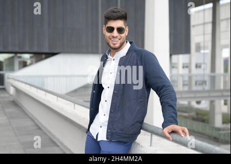 In der modernen Innenstadt steht ein junger, zwangloser Mann, der die Kamera anschaut und eine Sonnenbrille trägt. Hochwertige Fotos. Stockfoto