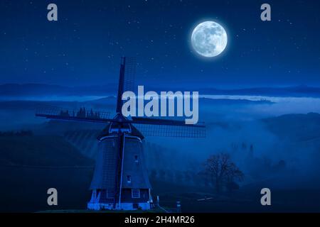 Nachtlandschaft mit Windmühle auf einem Hügel. Nebel im Tal und schöne Sterne am Himmel Stockfoto