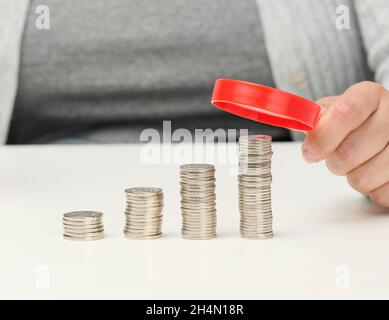 Frau sitzt an einem Tisch und hält eine rote Lupe über Stapel weißer Münzen. Das Konzept der Budgetplanung, Einsparungen. Investitionssuche Stockfoto