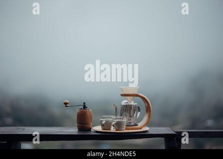 Ein Tropfkaffee-Set mit einem Stahlkessel, Glas und einer Kaffeemaschine mit Tropfwasser in minimalistischem Stil mit Nebel und Naturhintergrund. Stockfoto