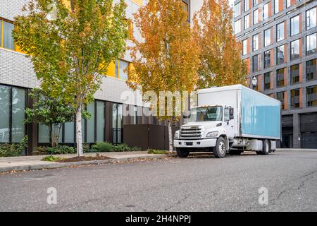 Weißer Mittelklasse-Umzugswagen Tagestab Sattelschlepper mit langem geräumigen Boxanhänger, der auf der städtischen Stadtstraße mit mehrstufigen Wohnungen steht Stockfoto