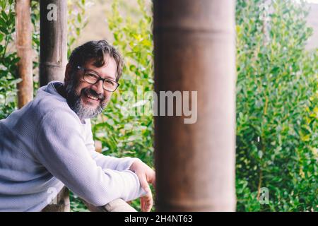 Fröhlicher junger, moderner reifer Mann lächelt und schaut auf die Kamera im Outdoor-Home-Portrait, das auf dem Balkon mit grünen Pflanzen im Hintergrund Holz steht. Happ Stockfoto