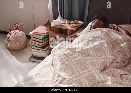 Afrikanische junge Frau liegt auf dem Bett und schaltet den Wecker aus, während sie morgens im Schlafzimmer aufwacht Stockfoto