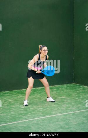 Positive Athletin mit lächelndem Schläger und wegblickend, während sie im Fitnessstudio mit grünen Wänden Padel spielt Stockfoto