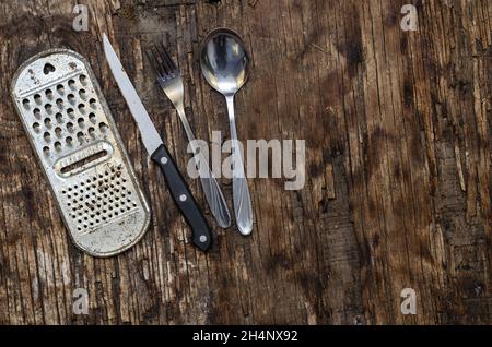 Alte rostige Küchenreibe, Messer, Löffel, Gabel gegen das verfaulende Brett. Küchenutensilien aus Metall. Draufsicht. Selektiver Fokus. Stockfoto