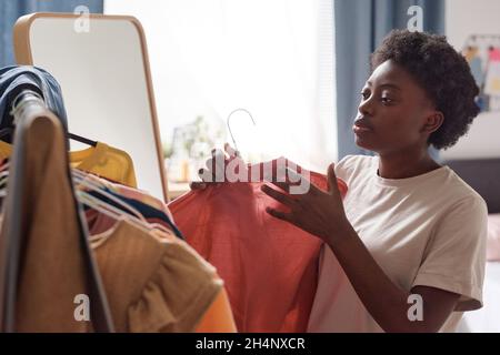 Afrikanische junge Frau, die Hemd auf dem Gestell wählt, um es zu tragen, während sie im Zimmer steht Stockfoto