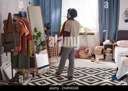 Rückansicht einer afrikanischen Frau, die sich für ein Kleid entschieden hat und auf den Spiegel schaute, während sie in ihrem Zimmer stand Stockfoto