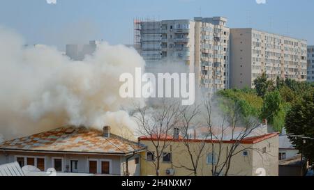 Flammen gehen aus brennendem Haus in der Nachbarschaft. Rauch, der aus dem Dach auf Feuer in der Stadtlandschaft auftaucht. Gefährliche Dämpfe und Smog, die durch eine Explosion aus dem zerstörten Gebäude entstehen Stockfoto