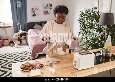 Afrikanische junge Frau, die während ihres Frühstücks im Zimmer heißen Kaffee in die Tasse gießt Stockfoto