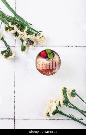 Tiramisu Dessert im Glas mit Beeren auf weißem Fliesenhintergrund mit Blumen Stockfoto