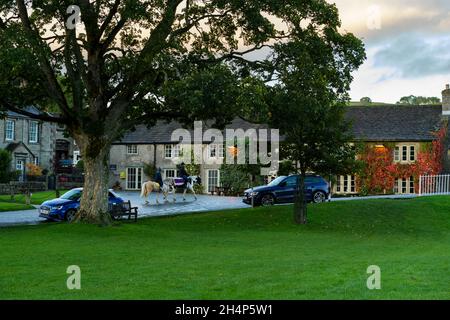 Idyllischer ländlicher Lebensstil - 2 Reiter auf der Straße bei Sonnenuntergang in der malerischen Burnsall (Hütten, alte Pension, Dorfgrün) - Yorkshire Dales, England, GB, UK. Stockfoto