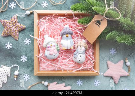 Geschenkbox mit Spielzeug auf grünem Hintergrund mit weihnachtlicher Dekoration. Es gibt drei Keramikspielzeug in einer Schachtel mit einem rosa Papierfüller, Schneemännern und einem Pinguin Stockfoto