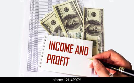 Dokumente, Dollar liegen auf weißem Hintergrund. Weibliche Hand schreibt auf einem Notizbuch EINKOMMEN UND GEWINN Stockfoto