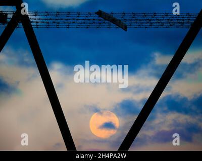 Ein Vollmond geht gerade unter, gesehen durch die rostenden Träger eines riesigen Masten auf einem Weizenfeld im Upper Radley Village. Die Sonne steht kurz vor dem Aufgehen Stockfoto