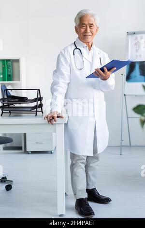 Lächelnder asiatischer Arzt hält Stift und Zwischenablage im Krankenhaus Stockfoto