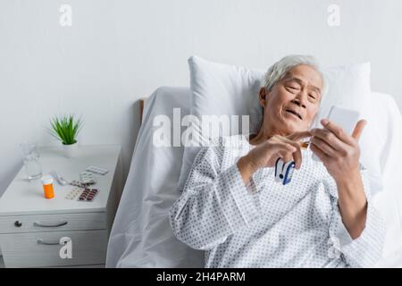 Älterer asiatischer Patient mit Pulsoximeter, der in der Klinik ein Smartphone am Bett in der Nähe von Pillen und Wasser verwendet Stockfoto