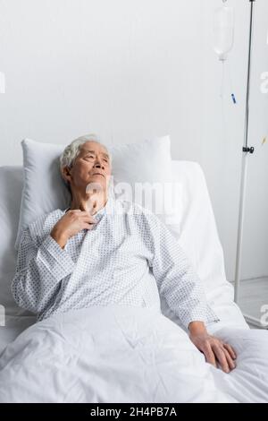 Kranker asiatischer Mann, der in der Nähe einer intravenösen Therapie auf der Krankenhausstation auf dem Bett liegt Stockfoto