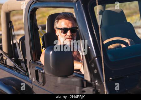 Porträt eines kaukasischen Mannes mit Sonnenbrille, der an einem sonnigen Tag am Meer im Auto sitzt Stockfoto